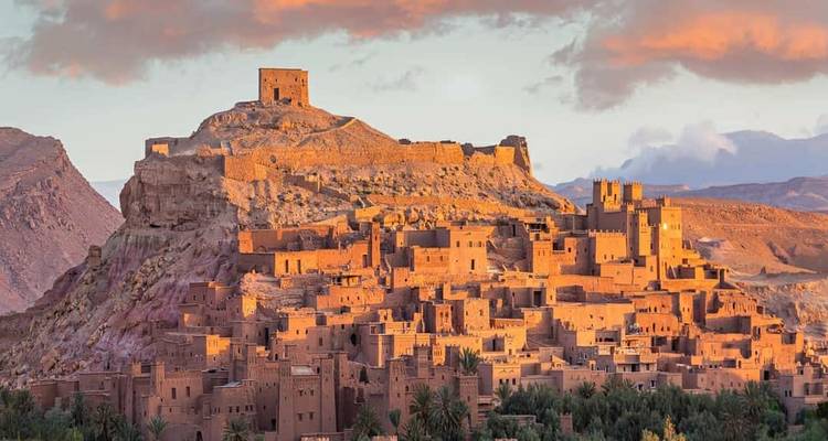 La kasbah en adobe d'Aït Benhaddou rougeoyant orange sous la lumière dramatique du coucher de soleil au sommet d'une colline rocheuse.