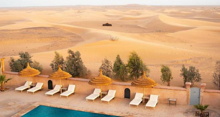 Station balnéaire du désert avec une piscine bleue et des rangées de transats donnant sur d'infinies dunes dorées.