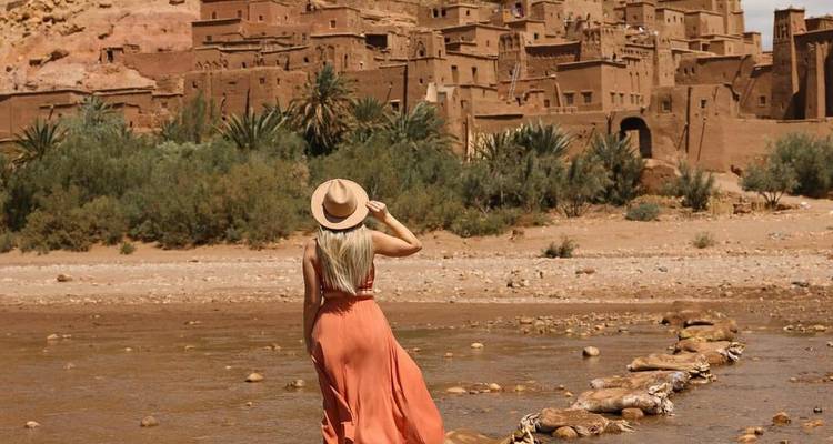 Voyageur en robe fluide contemple la forteresse en terre d'Aït Benhaddou au sommet d'une colline à travers un lit de rivière peu profond.