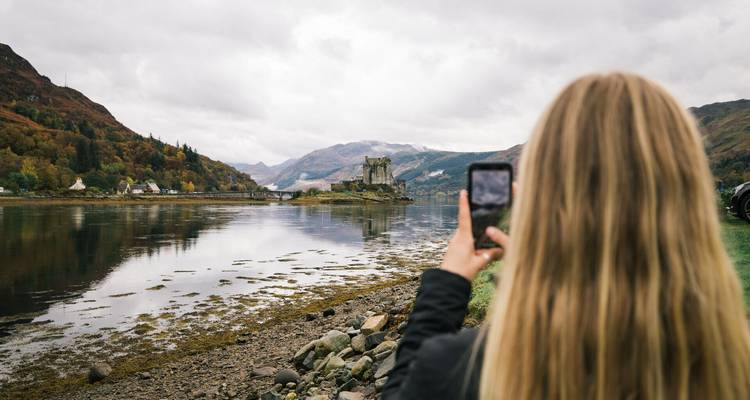 Une femme photographie un château sur un loch tranquille entouré de collines automnales.