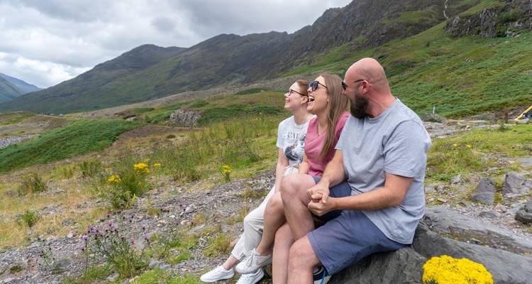 Trois voyageurs rient ensemble assis sur un rocher au milieu de fleurs sauvages dans les hautes terres vertes d'Écosse.