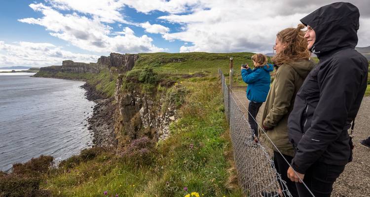 Des touristes emmitouflés dans des vestes admirent des falaises maritimes spectaculaires depuis derrière une clôture de sécurité par une journée venteuse.