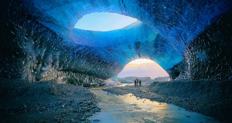 Abenteurer erkunden eine schimmernde blaue Eishöhle mit Sonnenlicht, das durch zwei Öffnungen hereinströmt.