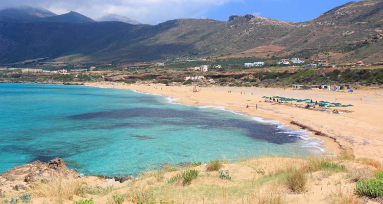 Wide sandy Cretan beach with turquoise water and distant mountains under a partly cloudy sky.