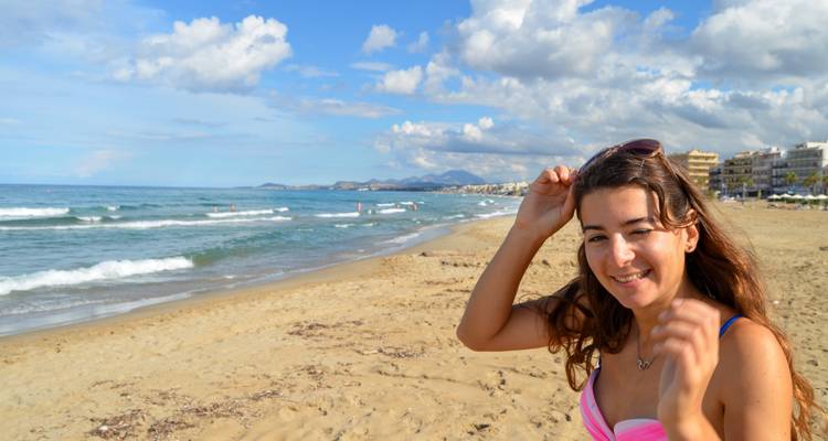 Young woman smiling on a sandy beach with gentle surf and distant shoreline.