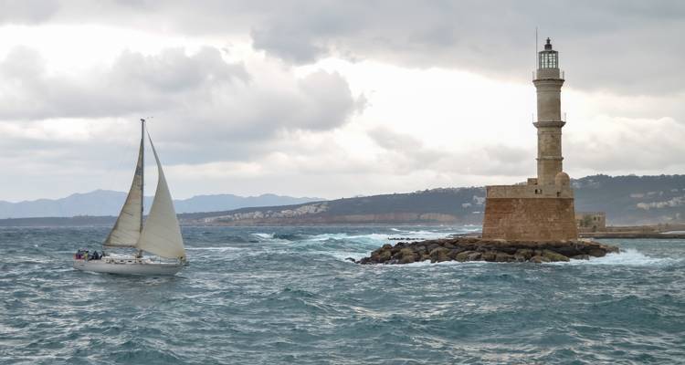 Sailing yacht passes the historic Venetian lighthouse at the entrance of Chania harbor on a windy day.