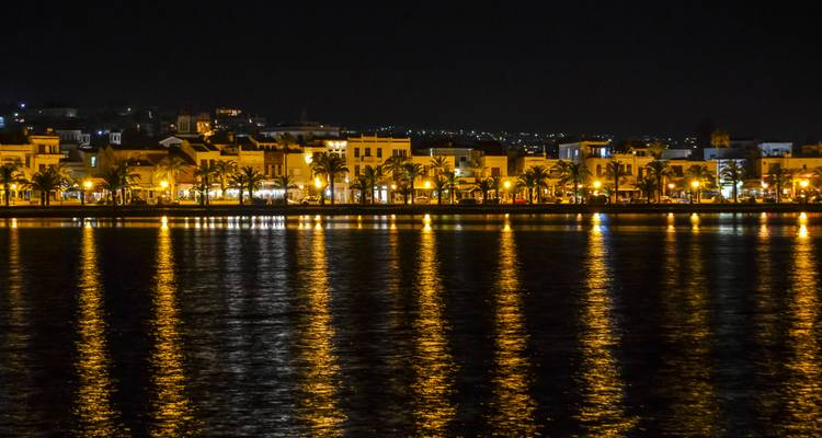Nighttime waterfront of Rethymnon with palm-lined promenade reflected in calm dark water.