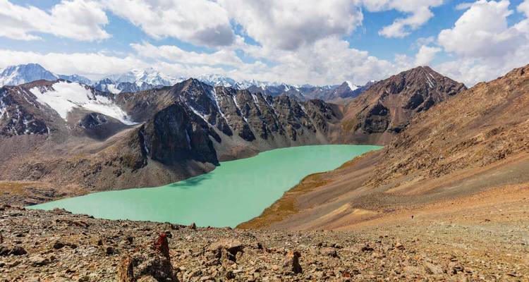 Lac alpin turquoise niché parmi des pics escarpés enneigés sous un ciel nuageux lumineux.