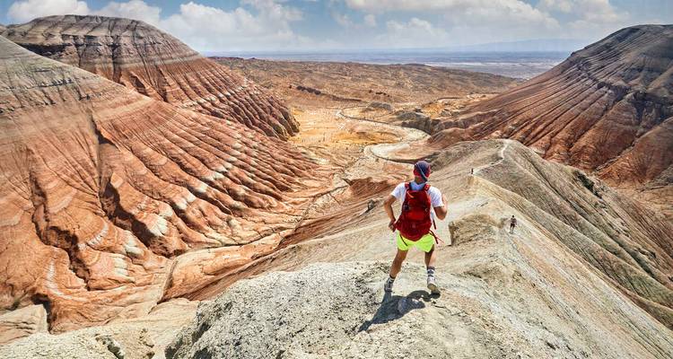 Randonneur en équipement coloré se tient sur une crête de canyon rayée rouge contemplant un vaste paysage désertique.