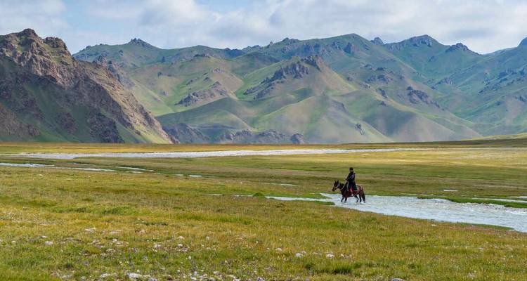 Un cavalier traverse une rivière peu profonde dans une vaste plaine verte bordée de montagnes ondulantes.