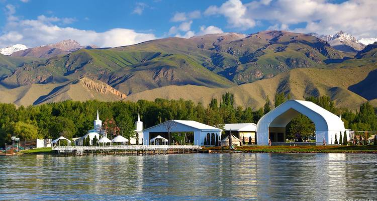 Des pavillons blancs au bord du lac se reflètent dans l'eau calme avec des montagnes saupoudrées de neige qui s'élèvent derrière.
