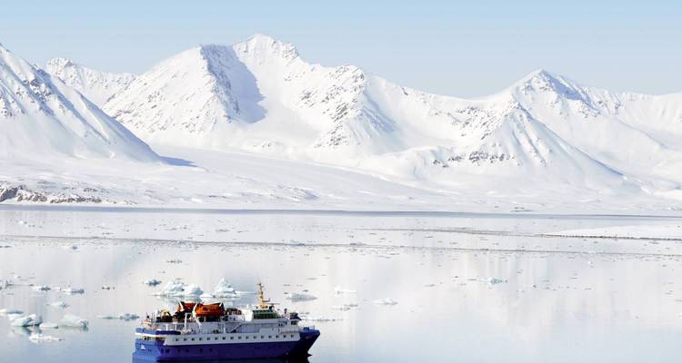 Expeditieschip varend in kalm ijzig water met torenhoge met sneeuw bedekte bergen erachter.