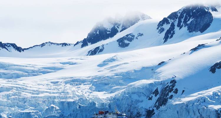 Blauw expeditievaartuig gedwarfd door dramatische gletsjers en ruige besneeuwde toppen.