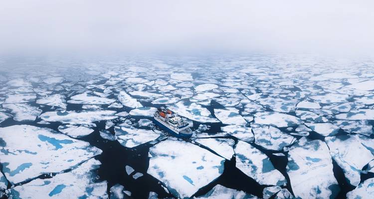 Luchtfoto van expeditie schip dat navigeert door een uitgestrekt mozaïek van zee-ijs onder mistige luchten.