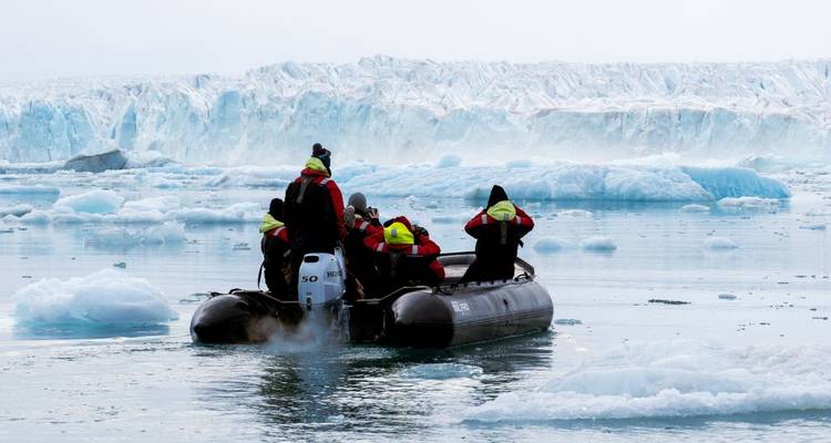 Zodiacboot met groepjes passagiers nadert torenhoge blauwe gletsjerwand te midden van drijvend ijs.