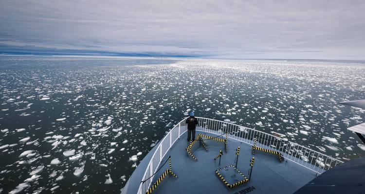 Persoon die aan de boeg van een schip staat en uitkijkt over uitgestrekt gebroken zee-ijs.