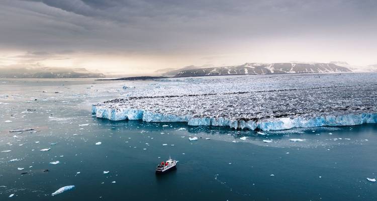 Kleine boot drijft nabij massieve gletsjerwand met bergen gehuld in wolken daarachter.