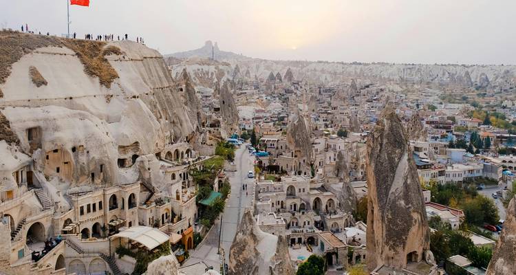 Weite Luftaufnahme der Stadt Göreme und der Feenkamin-Felsformationen in Kappadokien mit einer türkischen Flagge auf einem Hügel bei diesigem Sonnenuntergang