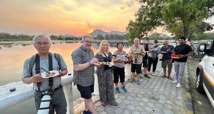 Toergroep geniet van snacks naast een rustig meer bij zonsondergang met gesilhouetteerde bergen.