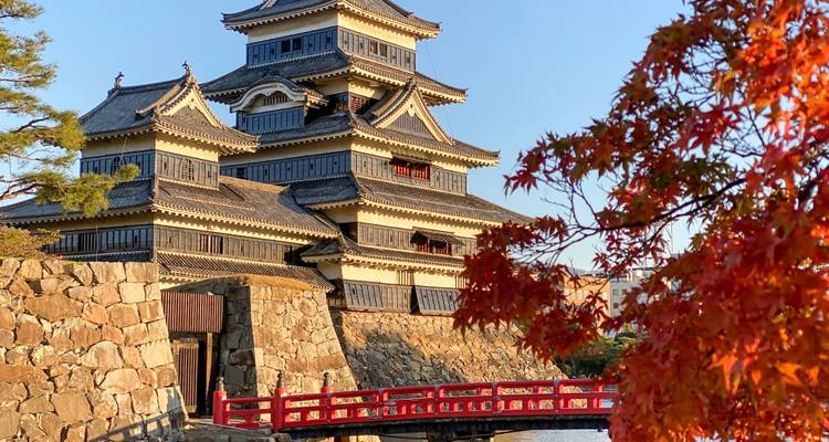 Un castillo japonés bien conservado con un puente rojo al frente enmarcado por hojas de arce ardientes de otoño bajo un cielo despejado.