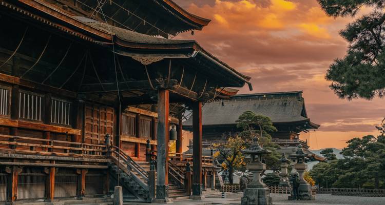 Los edificios históricos del templo de madera brillan cálidamente bajo un cielo al atardecer dramáticamente anaranjado y siluetas de pinos.