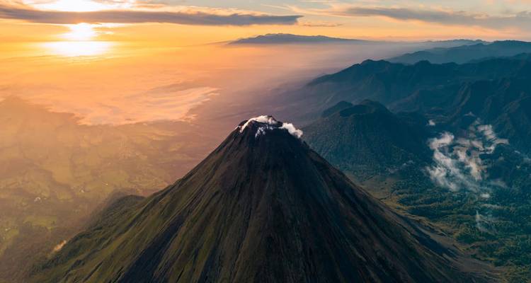 Dramática toma aérea de un volcán cónico humeante que se eleva sobre la selva tropical y el valle al amanecer.