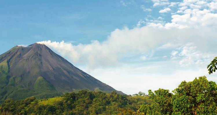 Vista lateral de un estratovolcán humeante que se alza sobre un exuberante bosque verde contra un cielo despejado.