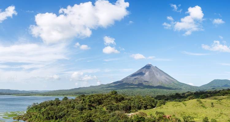 Panorama de un cono volcánico junto a un lago y selva tropical bajo nubes dispersas.