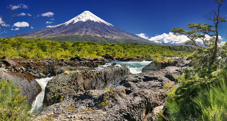 Volcán cubierto de nieve elevándose sobre un bosque esmeralda y rápidos de río torrencial bajo un cielo azul profundo
