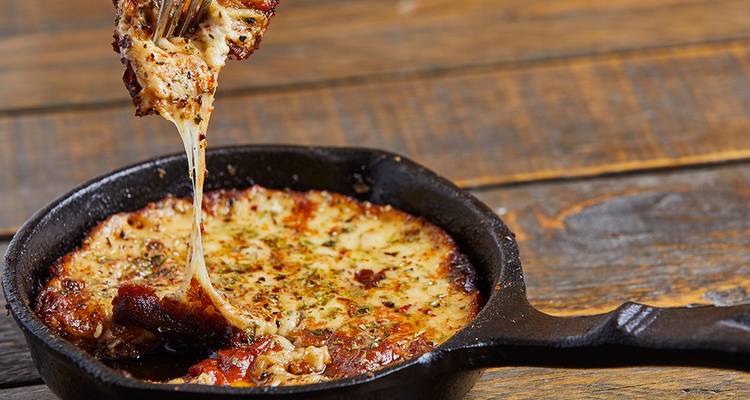 Primer plano de un plato de queso derretido chisporroteante siendo levantado de una pequeña sartén de hierro fundido