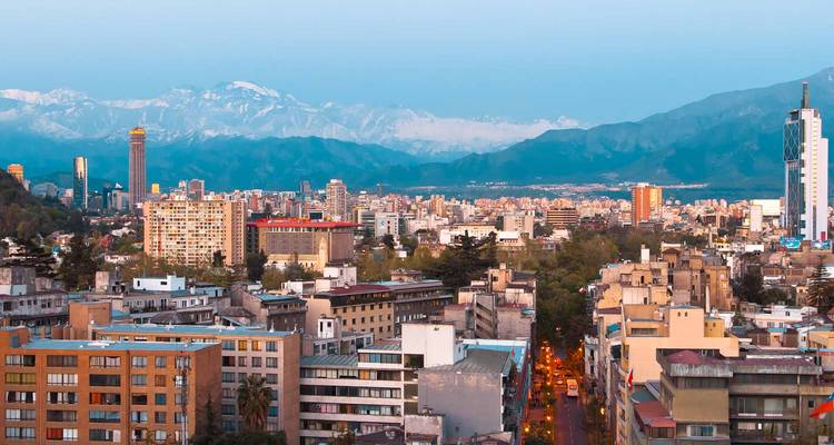Amplio paisaje urbano de Santiago con las montañas de los Andes cubiertas de nieve al fondo bajo un cielo azul despejado.