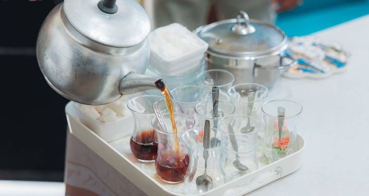 Nahaufnahme von türkischem Tee, der aus einem Metallkessel in schlanke Gläser auf einem Tablett gegossen wird.