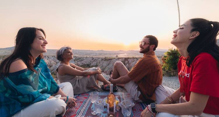 Vier Freunde entspannen sich bei Sonnenuntergang auf gemusterten Teppichen, teilen Getränke und haben die Täler Kappadokiens hinter sich ausgebreitet.