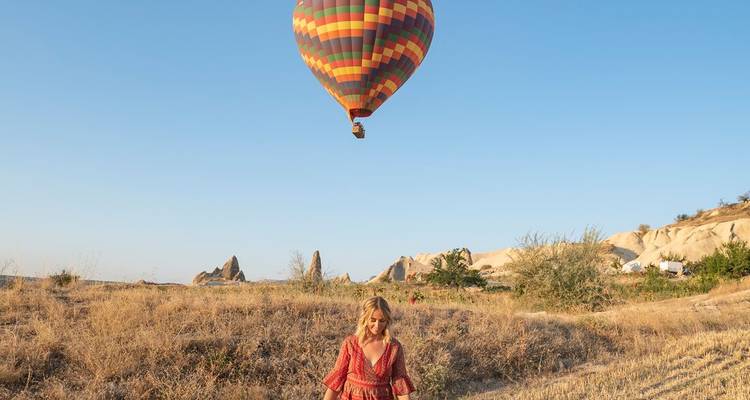 Eine Frau in einem roten Kleid steht in einem trockenen Feld, während ein bunter Heißluftballon über den Felsnadeln Kappadokiens schwebt.