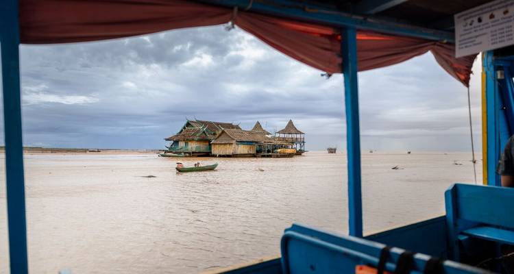 View from a blue wooden boat toward a floating village house on the expansive Tonle Sap Lake under grey skies.