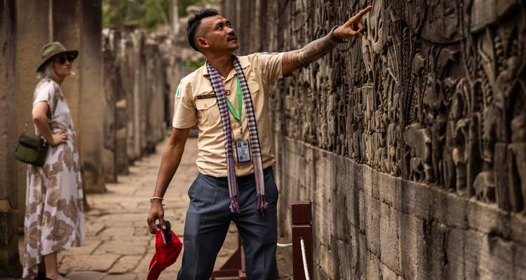 Local guide points out intricate bas-relief carvings on an ancient Angkor temple wall while a visitor observes.