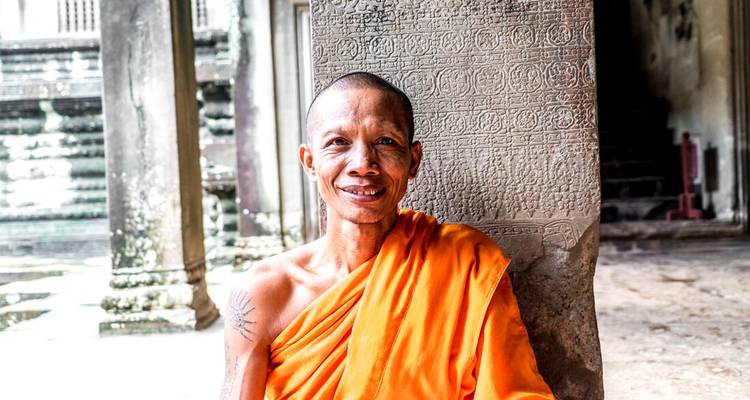 Portrait of a smiling Buddhist monk in orange robes leaning against a stone pillar engraved with Khmer script.