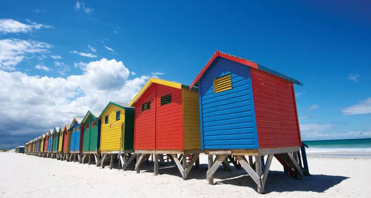 Fila de casetas de playa de madera pintadas de colores brillantes sobre arena blanca junto al mar turquesa y el cielo azul profundo.
