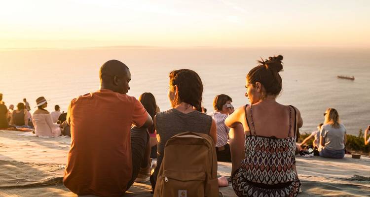 Los viajeros se sientan en una ladera al atardecer observando el tranquilo horizonte del océano volverse dorado.