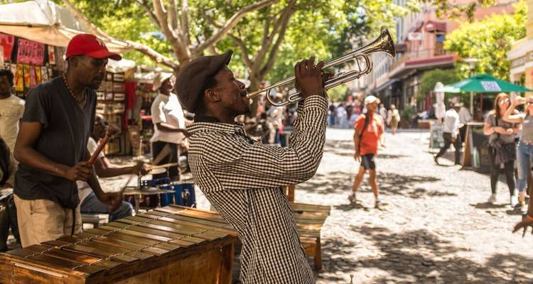 Músico callejero toca una trompeta enérgicamente en un mercado al aire libre, espectadores en el fondo.