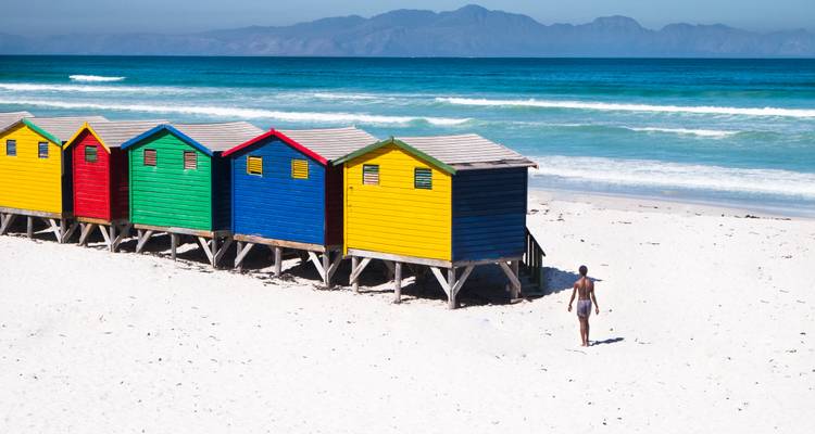 Fila de casetas de playa de colores brillantes sobre arena blanca con un bañista solitario caminando hacia las olas turquesas.