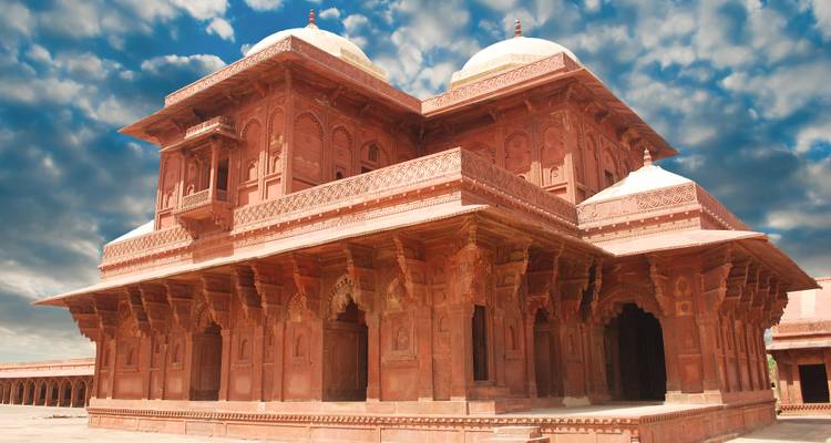 Palacio de arenisca roja con columnas talladas y cúpulas sobre un cielo dramático lleno de nubes.
