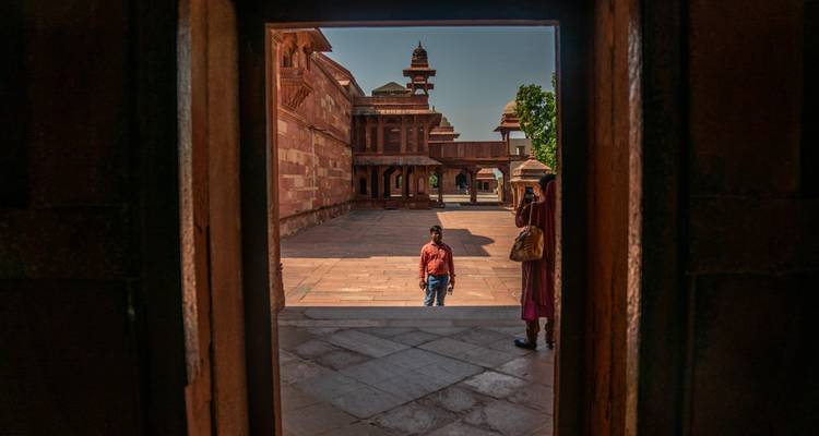 Vista a través de una entrada hacia un patio antiguo de arenisca donde dos viajeros se detienen bajo la luz del sol.