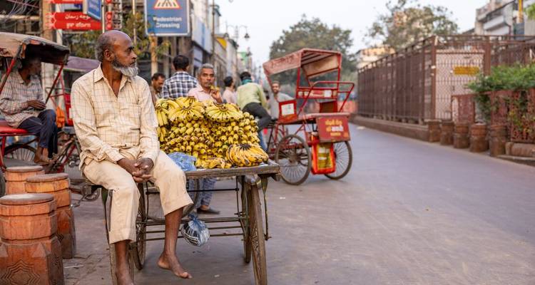 Barefoot street vendor sits on a cart piled high with bananas on a busy Delhi road.