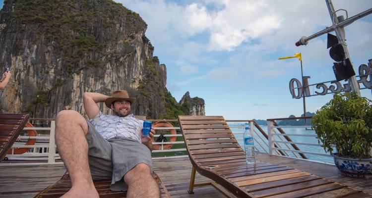 Man met een hoed ontspant op een ligstoel met een torenhoge kalkstenen klif en blauw water op de achtergrond