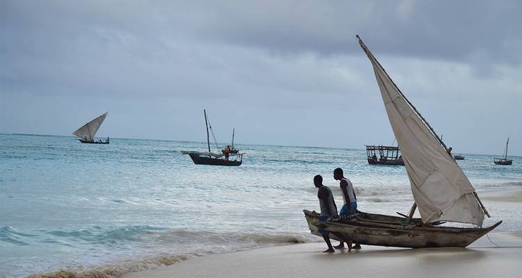 Two men maneuvering a small sailboat on a sandy beach with gentle surf.