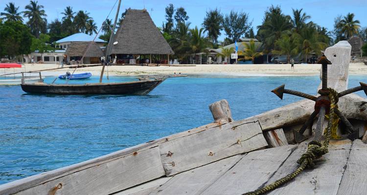 View from a wooden dhow’s bow toward a sandy shoreline with palm trees.