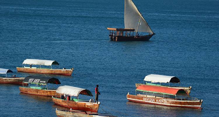 Row of small covered boats on deep blue water with a lone dhow sailing past.