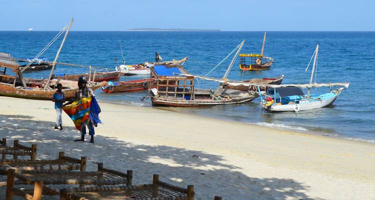 Beach scene with anchored wooden boats and vendors carrying colorful fabrics.