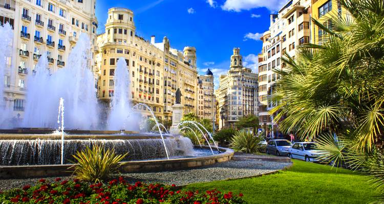 Großer Springbrunnen umgeben von Blumenbeeten inmitten prächtiger Gebäude aus dem frühen 20. Jahrhundert unter einem leuchtend blauen Himmel in Valencia.